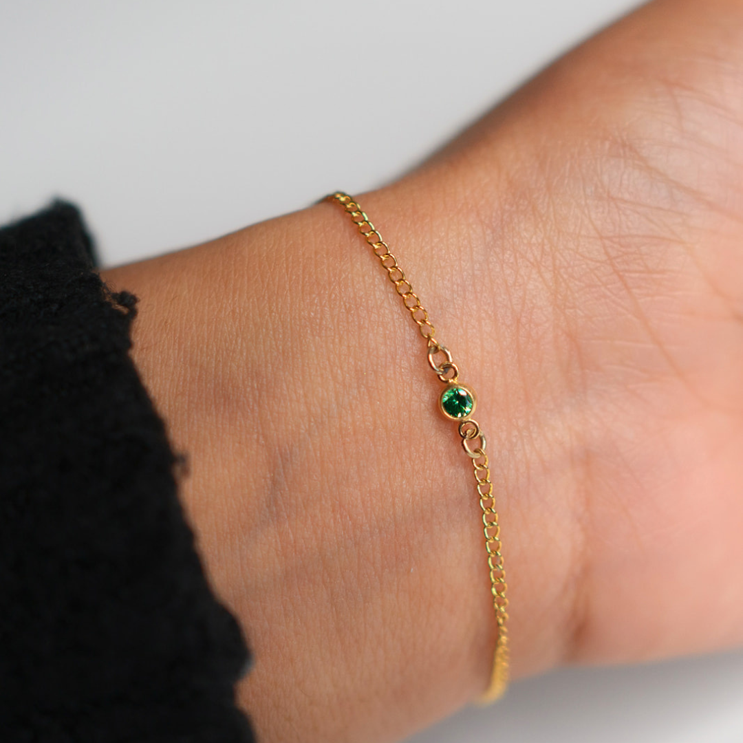 Close-up of a delicate gold bracelet with a small green gemstone on a wrist, highlighting luxury permanent jewellery, custom permanent jewellery styles, and exclusive permanent jewellery for special occasions.