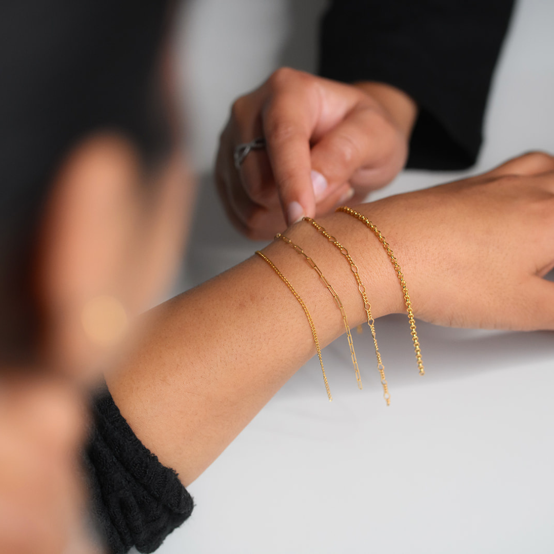 A woman wearing three different styles of gold bracelets on her wrist, representing personalised permanent jewellery, gold permanent jewellery, and handmade permanent jewellery from our Birmingham studio.